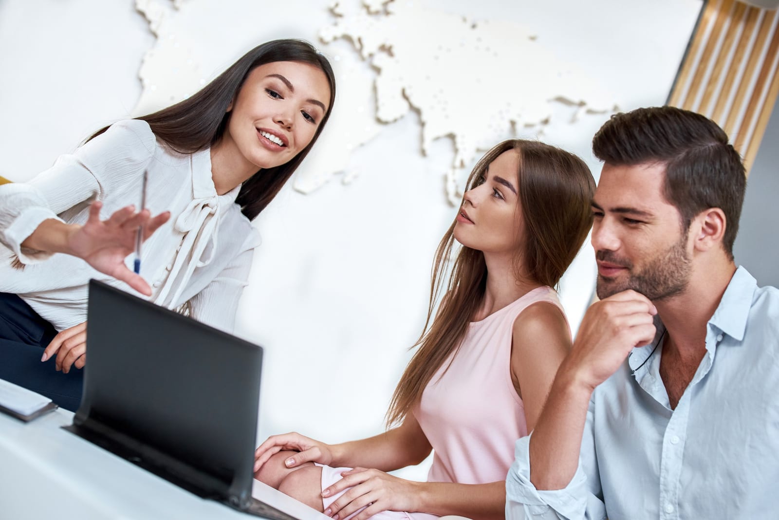 Atendente mostra opções no notebook para um casal em uma sala com mapa-múndi na parede